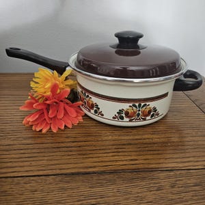 May include: A vintage enamel saucepan with a dark brown lid and handle. The off-white body features a floral design in shades of brown, orange, and green. A cluster of orange and yellow flowers sits beside the pan on a wooden surface.