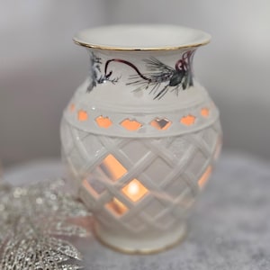 May include: A white ceramic vase with a lattice design and a gold rim. The vase has a floral pattern and diamond-shaped cutouts that emit a warm glow from the candle inside. A decorative piece for home decor.