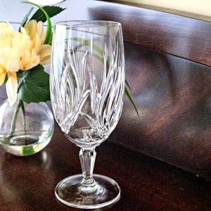 May include: A clear crystal champagne flute with a decorative etched design. The glass has a long stem and a wide base. A small glass vase with yellow flowers is in the background.