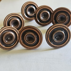 May include: A collection of eight antique-style cabinet knobs. Each knob features a dark bronze center surrounded by concentric rings and a beaded border, with a copper-colored outer rim. The knobs are circular and have a classic, decorative design.