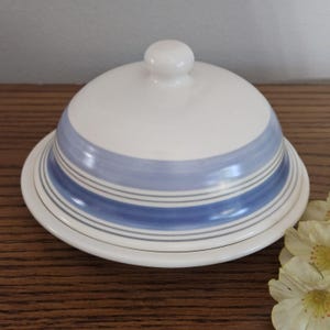 May include: A white ceramic butter dish with a domed lid. The lid and base are decorated with blue and gray horizontal stripes. A small, round knob sits atop the lid. The dish is on a wooden surface.