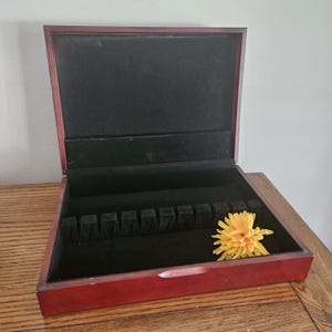 May include: Open, rectangular wooden silverware storage box with a dark red finish and black velvet interior. The box has slots for utensils and a yellow flower rests inside.