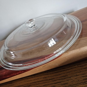 May include: A clear glass lid with a small knob handle. The lid is oval-shaped and sits on a wooden surface. The glass is transparent, allowing a view of the surface below. The lid is designed for cookware.