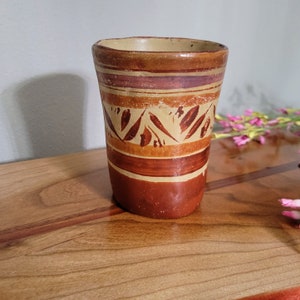 May include: A cylindrical earthenware cup featuring a hand-painted design with horizontal bands in shades of terracotta, cream, and dark brown. The central section displays a repeating pattern of stylized leaf shapes in a dark brown pigment.