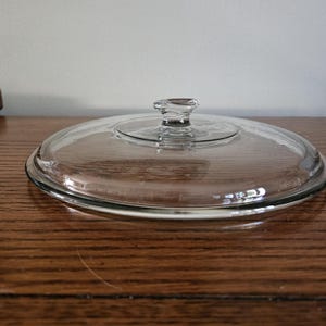 May include: A clear glass lid with a small knob handle. The lid is round and appears to be for a pot or pan. The glass is transparent, allowing a view of the contents below. The lid is resting on a wooden surface.