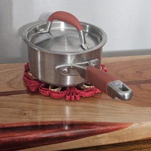 May include: A stainless steel saucepan with a lid and a red handle, resting on a red and gold woven trivet. The handle on the lid is also red. The pot is on a wooden surface.