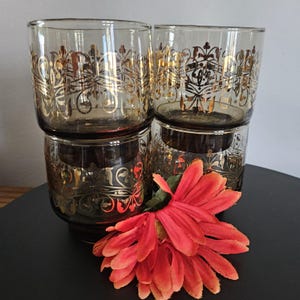 May include: Set of four amber glass tumblers with gold leaf designs. The glasses are stacked on top of each other and a red flower is in front of them.