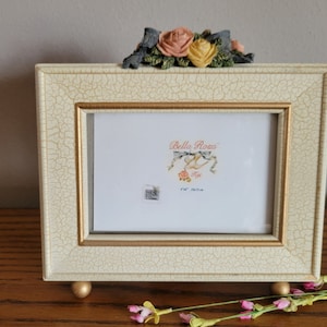 May include: A white picture frame with a distressed finish and gold accents. The frame has a floral design on the top and a small label that says "Bella Rosa" with a bird and the text "4" x "6" 10.16cm x 15.24cm".