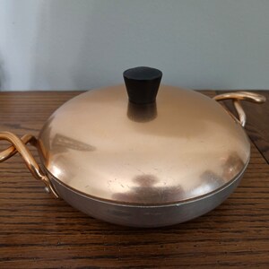 May include: A round, shallow cooking pan with a polished copper lid and a black knob handle. The pan features two curved, metallic gold-toned side handles and a silver-colored base, presenting a classic design for kitchen stovetop use.