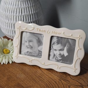 May include: Cream-colored ceramic double photo frame with the words "Then & Now" in raised lettering. The frame holds two black and white photos and features a decorative beaded border. A white vase and a daisy are in the background.