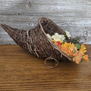 May include: A brown woven wicker cornucopia basket filled with artificial flowers in shades of white, yellow, and orange. The horn-shaped basket features a natural twig texture and a small circular loop at the base, holding a cluster of silk-like blooms.