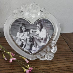 May include: A heart-shaped clear glass picture frame featuring a frosted rose and leaf design at the top and bottom. The frame holds a black and white photograph of a family of three sitting together on a beach, with a smooth, polished finish.