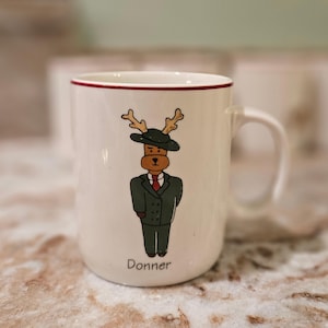 May include: White ceramic mug with a red rim and a handle. The mug features a cartoon illustration of a reindeer wearing a green suit, a black hat, and a red tie. The name "Donner" is printed at the bottom.