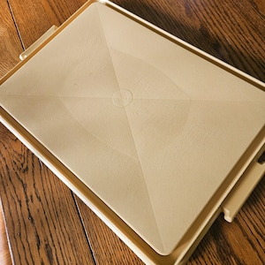 May include: A beige plastic serving tray with a textured surface and geometric design. The tray has a rectangular shape with a raised edge and handles on the sides. The tray is sitting on a wooden surface.