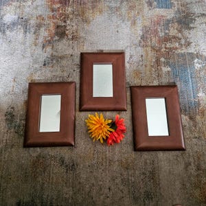 May include: Three rectangular brown wooden frames with mirrors inside, arranged on a patterned surface. Two artificial flowers, one orange and one red, are placed between the frames. The frames are empty and ready for display.
