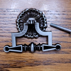 May include: A decorative silver-toned drawer pull with a floral design and a matching screw. The pull has a rectangular handle with a central, ornate design. The hardware is set against a wooden surface.