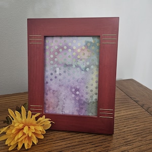 May include: A rectangular, burgundy-colored picture frame with a watercolor print. The frame has thin, horizontal gold-colored accents on the sides. A yellow flower rests on the wooden surface in front of the frame.