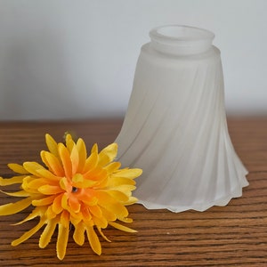 May include: A frosted glass lampshade with a spiral design, next to a bright yellow artificial flower. The lampshade has a flared shape and sits on a wooden surface.