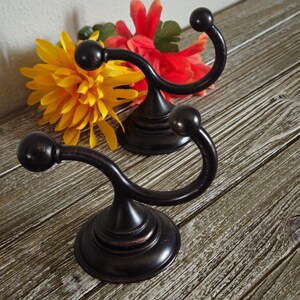 May include: Two dark bronze-toned metal wall hooks with a curved design and spherical ends. Each hook features a circular base and a smooth, polished finish. The hooks are positioned in front of bright yellow and orange artificial flower petals.