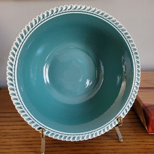 May include: A teal ceramic bowl with a white rope-like border. The bowl is displayed on a gold-colored stand. The interior of the bowl is a solid teal color. The rim of the bowl has a white, textured design.