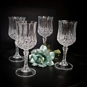 May include: Four clear crystal wine glasses with diamond-cut detailing. The glasses have a tulip shape and sit on a round base. A light blue flower is placed near the glasses against a black background.