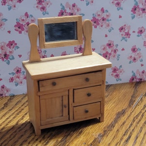 May include: A miniature wooden dresser with a mirror, featuring a light brown finish. The dresser has three drawers, a cabinet door, and two decorative posts on either side of the mirror. The background has a floral pattern.