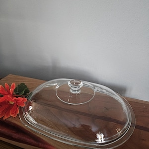 May include: A clear, oval-shaped glass casserole dish lid with a small, round handle. The lid sits on a wooden surface, with a red flower and green leaves in the corner. The lid is approximately 12 inches long.