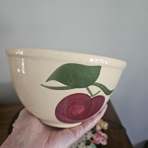 May include: A cream-colored ceramic mixing bowl featuring a hand-painted design of a dark red cherry with a green leaf and stem on the exterior. The bowl has a smooth, rounded rim and a tapered base, showcasing a glossy finish throughout the surface.