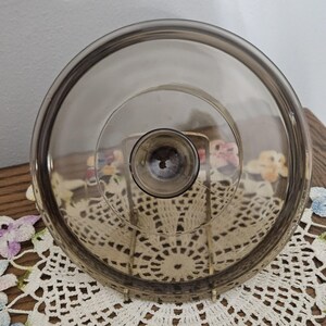 May include: A circular, smoke-tinted glass lid featuring a smooth, transparent surface and a centered, raised knob handle. The lid has a slightly elevated inner rim and a wider, flat outer edge, providing a clear view through the dark glass.