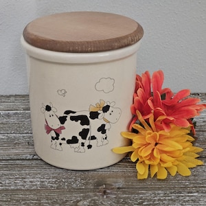 May include: A white ceramic jar with a wooden lid, featuring a black and white cow design with red and yellow accents. The jar has a whimsical illustration of two cows with a cloud and a bird in the background.
