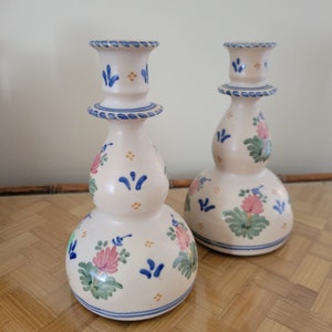 May include: Two off-white ceramic candle holders with a bulbous shape. Each is decorated with blue and pink floral designs, blue accents, and a blue rim. The candle holders are displayed on a light brown surface.