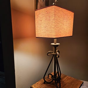 May include: A decorative table lamp with a square lampshade in a warm orange hue. The lampshade is trimmed with a dark brown border. The lamp has a dark metal base with a tripod design and decorative loops. The lamp sits on a wooden table.