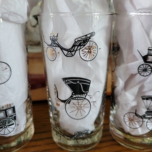 May include: Clear glass tumblers with black and gold carriage designs. The vintage-style glasses feature various carriage illustrations, with gold accents on the wheels. The glasses are filled with white tissue paper.