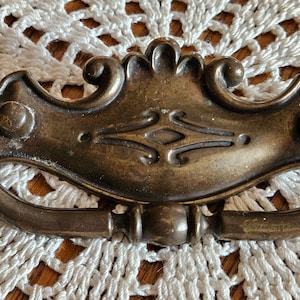 May include: Antique brass drawer pull with a decorative design. The pull features a curved handle and ornate detailing, including a central diamond shape and scrollwork. The hardware is likely for a cabinet or dresser.