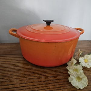 May include: An orange and red enameled cast iron Dutch oven with a lid and two handles. The lid has a black knob. The pot is sitting on a wooden surface with white flowers.