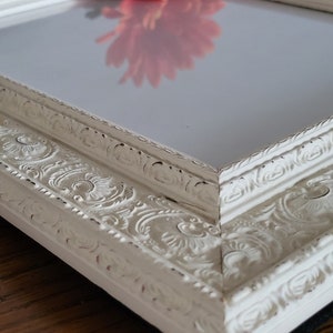 May include: Close-up of a white, ornate picture frame with a distressed finish. The frame features detailed floral and scrollwork carvings. A red flower is reflected in the glass. The frame is on a wooden surface.