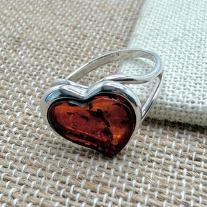 Może przedstawiać: Srebrny pierścionek z bursztynem w kształcie serca. Obręcz pierścionka otacza bursztynowe serce, tworząc unikalny design. Bursztyn ma ciepły, czerwonobrązowy kolor.