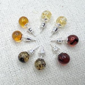 May include: A collection of stud earrings with silver posts and various colored, round, translucent beads. The colors include amber, yellow, and dark red. The earrings are arranged in a circular pattern on a textured, off-white surface.
