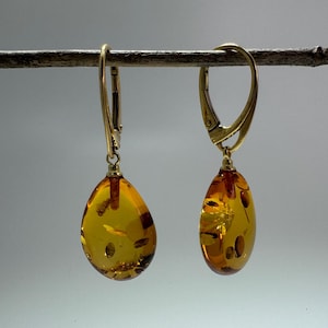 May include: A pair of gold-colored drop earrings with teardrop-shaped amber stones. The amber stones are a translucent yellow-orange color with visible inclusions. The earrings are hanging from a thin, brown branch against a light background.