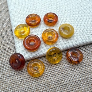 Op de afbeelding: Een verzameling amberkleurige, donutvormige kralen in verschillende tinten geel, oranje en bruin. De kralen zijn gerangschikt op een witte stof en een getextureerd beige oppervlak. De kralen hebben een gat in het midden.