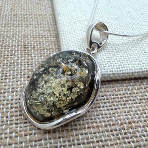 May include: A silver pendant necklace featuring a large, oval-shaped amber stone with gold inclusions. The amber is set in a polished silver bezel and hangs from a delicate silver chain. The pendant has a unique, organic appearance.