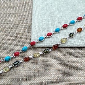 May include: Two silver bracelets with oval-shaped gemstones. One bracelet features turquoise and red stones, while the other has amber, yellow, and green stones. The bracelets are displayed on a textured, neutral-toned surface.