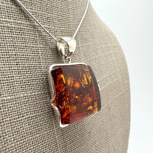 May include: A silver necklace featuring a large, rectangular amber pendant. The amber is a rich, warm brown color with visible inclusions. The pendant is set in a silver frame and hangs from a delicate silver chain.
