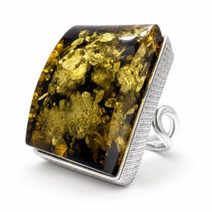 Könnte beinhalten: Ein Ring mit einem großen, rechteckigen, polierten Bernstein mit dunkelgrünen und goldenen Einschlüssen. Der Stein ist in eine texturierte, silberfarbene Metallfassung mit Kreuzschraffur an den Seiten und einer gedrehten Ringschiene gefasst.