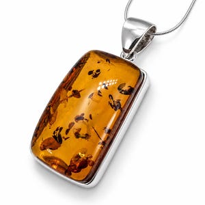 Op de afbeelding: Een rechthoekige hanger met een gepolijste, honingkleurige barnsteen met zichtbare donkere organische insluitingen, gezet in een glad zilveren montuur. De hanger hangt aan een fijne zilveren ketting met een glanzende metalen afwerking.