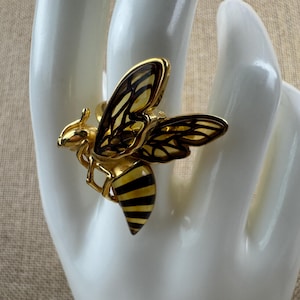 Op de afbeelding: Een goudkleurige bijenring met zwart-gele strepen op het achterlijf. De vleugels zijn doorschijnend met een bruin patroon. De ring is geplaatst op een witte hand.