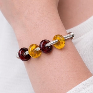 May include: A silver bracelet with four glass beads in shades of amber and burgundy. The beads are round and have a textured appearance. The bracelet is worn on a wrist.