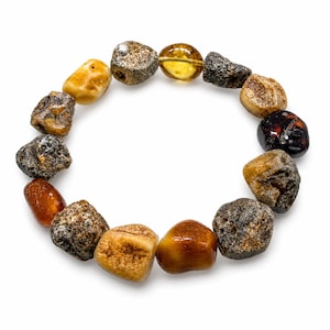 May include: A bracelet composed of various amber stones in shades of yellow, brown, and black. The stones are irregularly shaped and polished, with a natural, organic appearance. The bracelet is set against a white background.