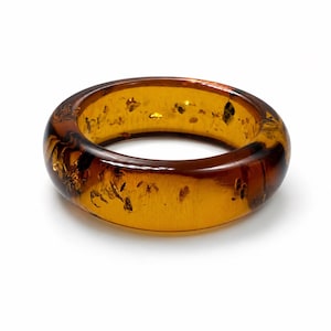 May include: A smooth, rounded bangle bracelet crafted from translucent amber-coloured material. The circular band features a polished finish with visible dark, organic inclusions suspended throughout the warm, golden-brown resin structure.