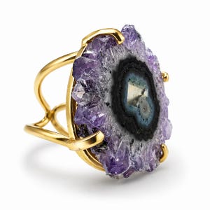 May include: A gold-toned cuff bracelet featuring a large, polished amethyst geode. The geode has a central dark eye with concentric rings of black, blue, and white, surrounded by a rough, crystalline purple exterior. The bracelet's design is open and airy.
